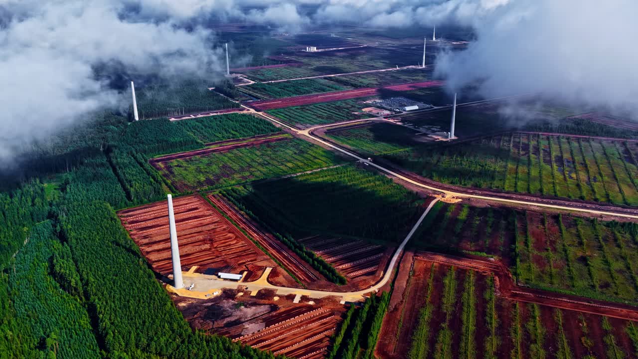 Drone flight over a developing renewable energy park in rural Latvia featuring newly built white turbine towers among green pine forests and vast agricultural farmlands