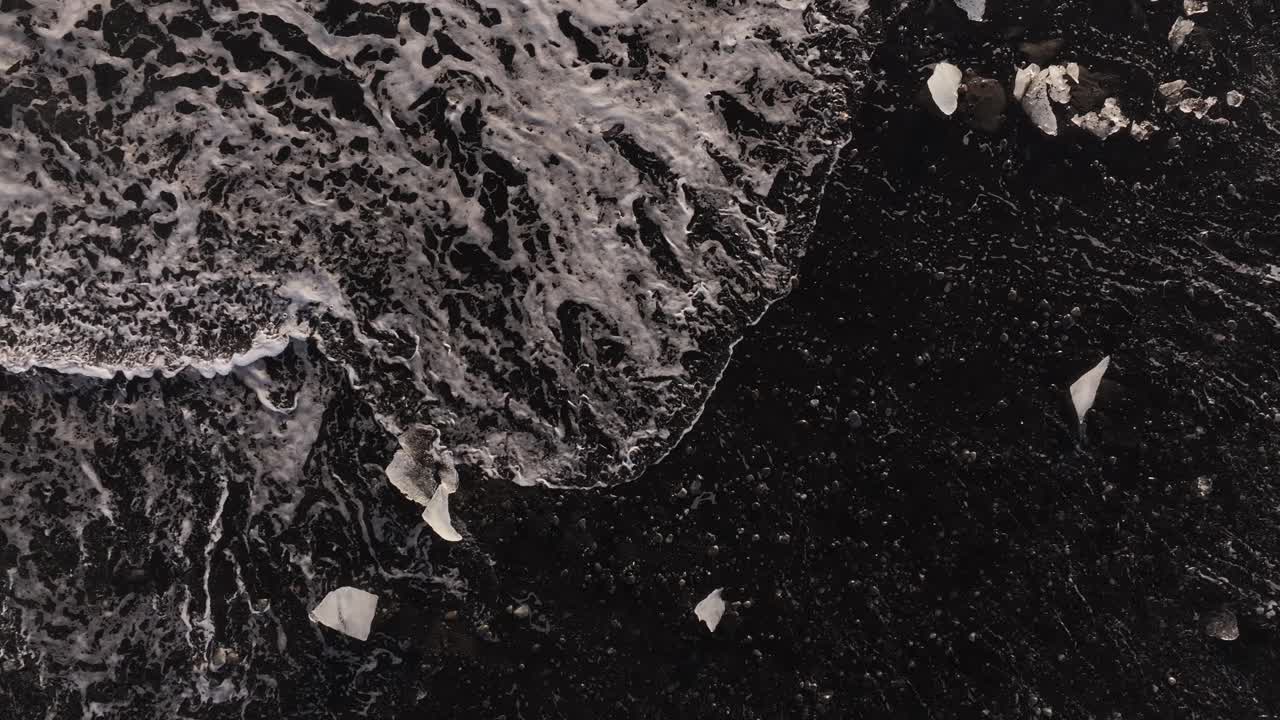 Top-down view of foamy waves crashing over black sand and scattered ice pieces at Diamond Beach, Jökulsárlón, Iceland.