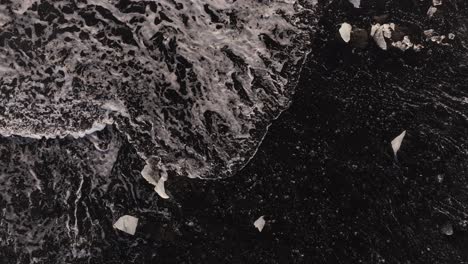 top-down view of foamy waves crashing over black sand and scattered ice pieces at diamond beach, jökulsárlón, iceland.