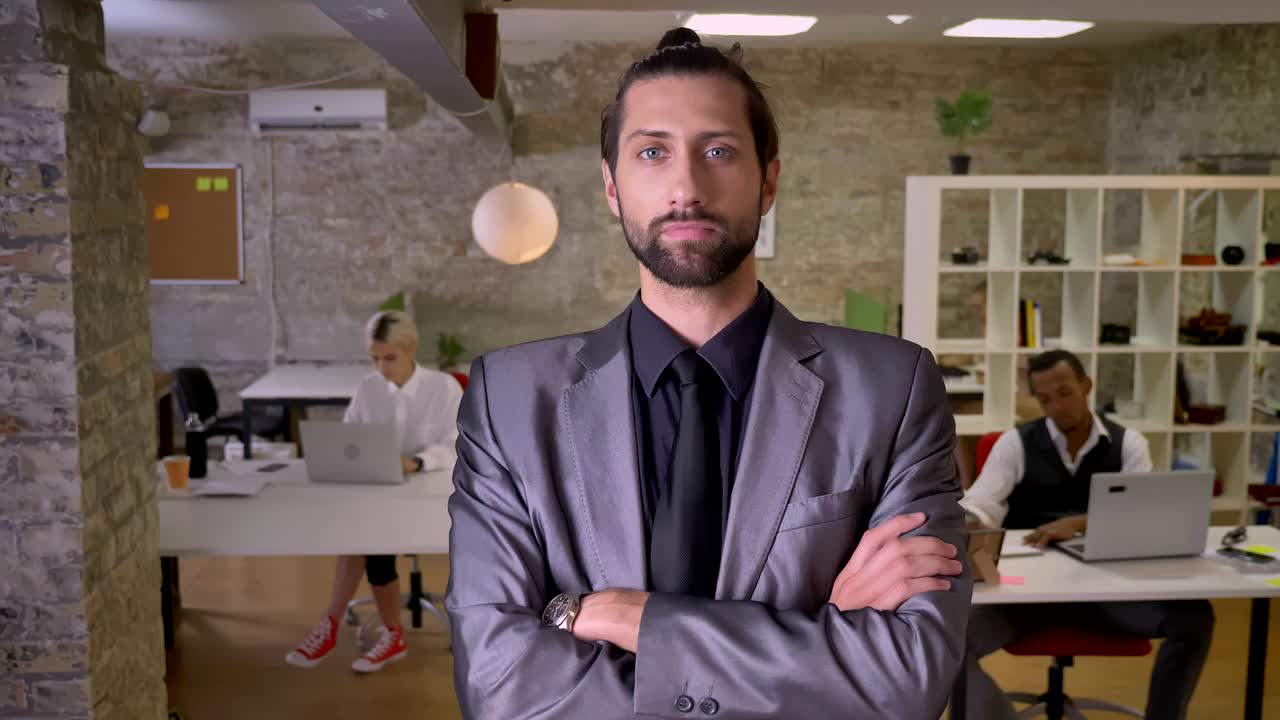 Handsome confident businessman with beard is standing with arms crossed and watching at camera in office, colleagues are working with laptops, work concept