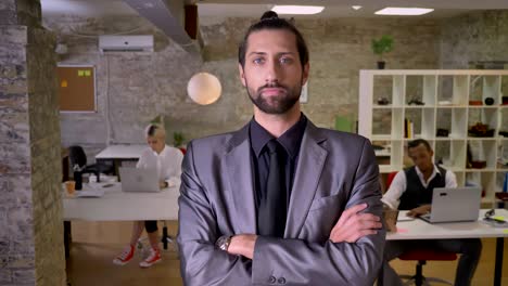 handsome confident businessman with beard is standing with arms crossed and watching at camera in office, colleagues are working with laptops, work concept