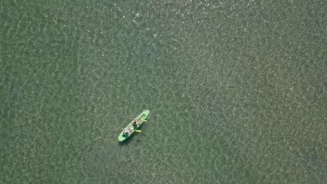 two people in sit-on-top kayak paddle in shallow green water, aerial