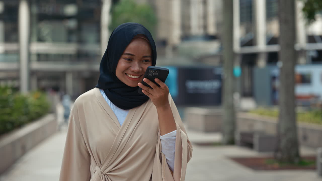 Young woman in hijab smiling while talking on smartphone outdoors in a city