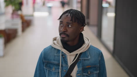close up view of young man wearing denim jacket layered over hoodie walking through bright mall corridor glancing sideways with thoughtful expression blurred storefronts and ceiling lights behind him