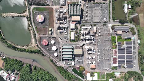 drone shot of wastewater treatment plant showing tanks, industrial buildings, filtration units, parking lots, nearby river, farmlands, and green zones on a clear sunny day