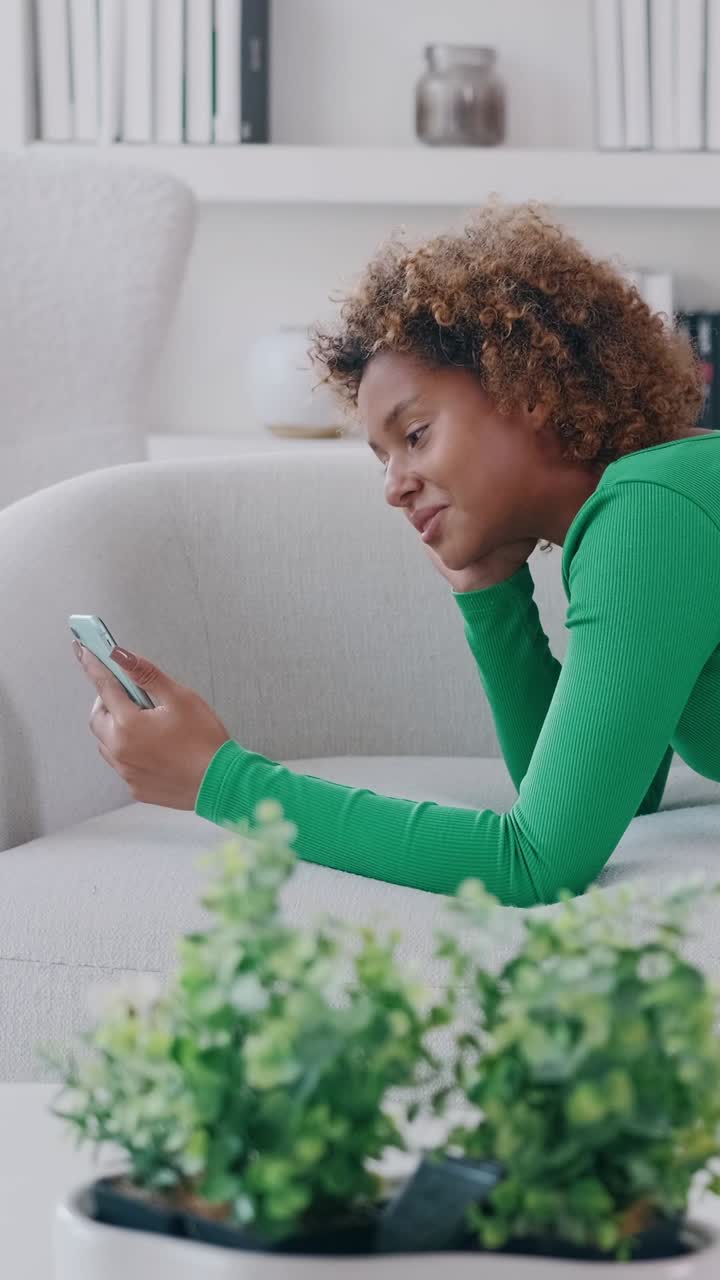 Young woman relaxed at home lying on a sofa using a smartphone and smiling