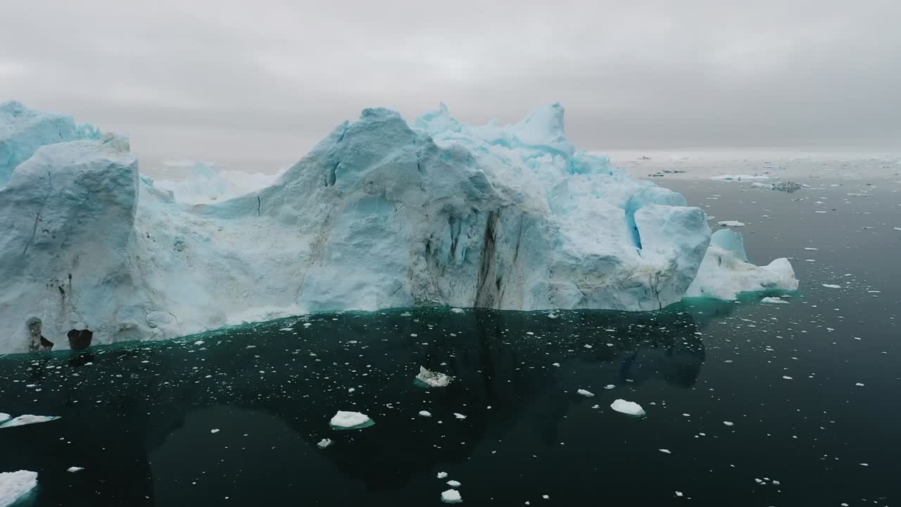Aerial drone view of a massive iceberg drifting in the dark Arctic sea near Greenland, showing stunning textures, cracks, and shades of blue in a frozen polar landscape