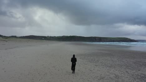 Vista-Trasera-Del-Hombre-De-Negro-Caminando-En-La-Playa-De-Xago-En-Asturias-España-En-Un-Día-Nublado---Tiro-De-Seguimiento-De-Drones