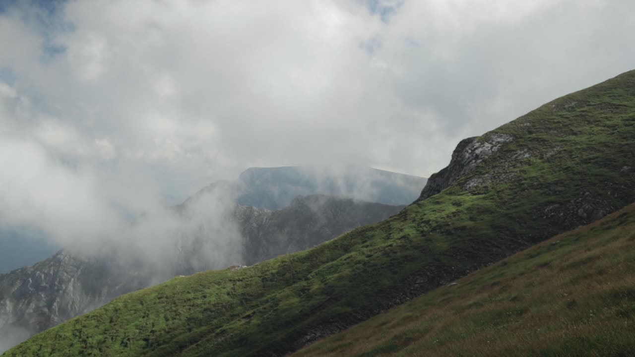 panorámica de izquierda a derecha de una montaña empinada con nubes bajas y picos de montaña en el fondo