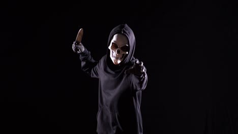 hombre aterrador en la sudadera con capucha usando una máscara de halloween sosteniendo un cuchillo y corriendo sobre el fondo negro