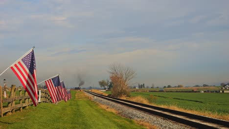 una vista de una sola vía ferroviaria, con una valla con la bandera de américa en ella, agitando suavemente en el viento a medida que se acerca un tren de vapor, en un soleado día de otoño