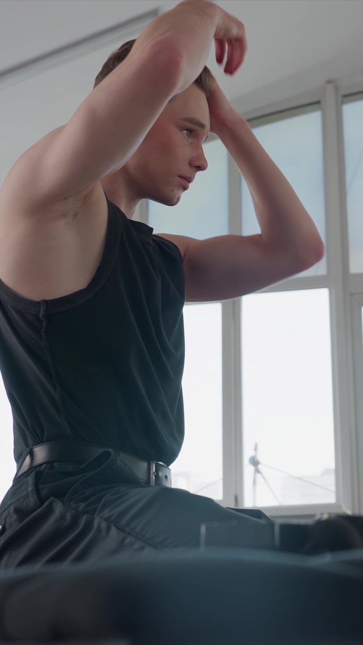 Young man in black sleeveless top and dark pants performs. Man transitions to stretching on exercise machine raising. Scene remains consistent throughout with bright natural light
