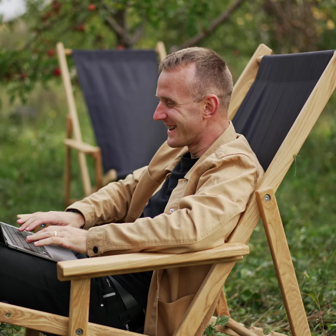 Emotional talk through the video chat on laptop. Man holding computer on his laps sitting in a garden chair outdoors. Apple garden blurred backdrop