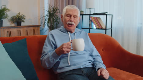 Attractive-smiling-senior-old-grandfather-man-drinking-a-cup-of-coffee-or-herbal-tea-at-home-couch