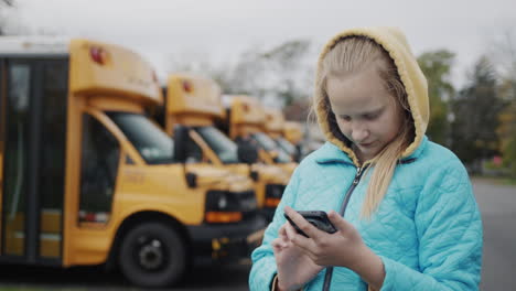 un escolar usa un teléfono inteligente, de pie contra el fondo de los autobuses escolares amarillos