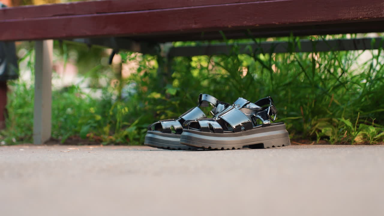 공원 벤치 가까이, 덩그러니 놓인 샌들. 나무 벤치 아래 콘크리트 위에 놓인 반짝이는 검은색 가죽 샌들, 녹색 잎과 이끼 배경, 부드러운 아침 햇살, 얕은 피사계 심도, 텍스처