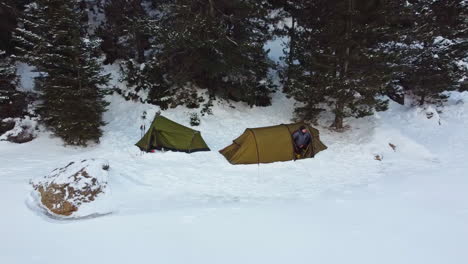 pull back aerial shot from a camping tents,over a frozen lake, covered of snow,at the background a mountain full of snow, revealing forest, while men getting the heads out of tents to see the scenery