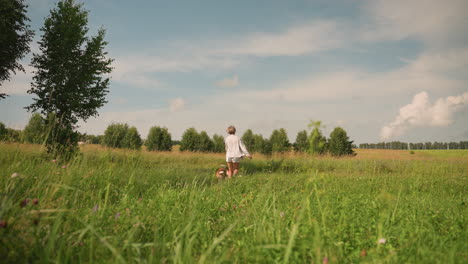 back view of woman running joyfully with two dogs on leash through grassy field under clear sky, showcasing fun outdoor interaction with pets, background features lush greenery and trees