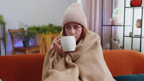 Sick-redhead-woman-wear-hat-wrapped-in-plaid-sit-alone-shivering-from-cold-on-sofa-drinking-hot-tea