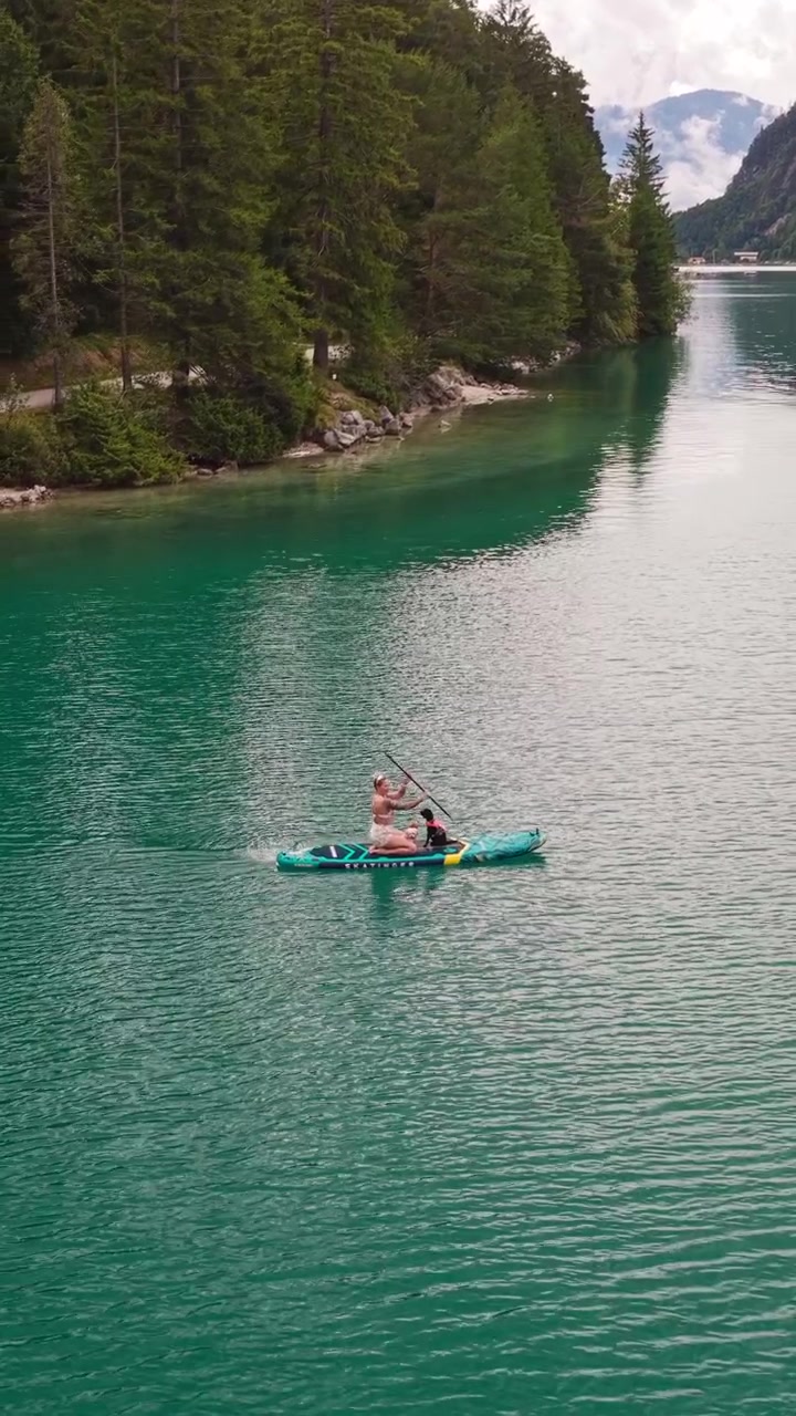 Una donna siede su una tavola da stand-up paddle con cani a bordo che pagaiamo sul lago Achensee in Tirolo, Austria. Ripresa verticale, aerea.