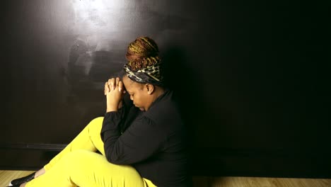 sideview of black woman sitting on floor against black wall praying to god