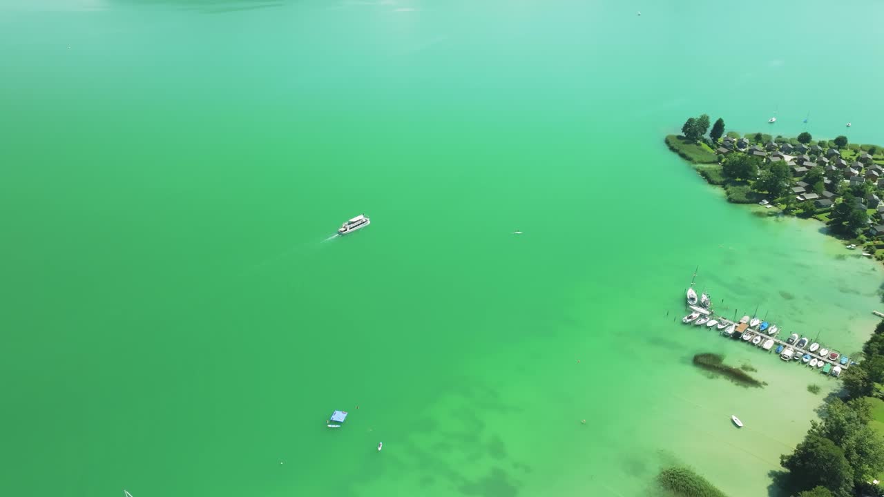 Aerial view of Mondsee's serene emerald waters, revealing a luxurious boat crossign the lake diagonally swimming close to a pier and a holiday village