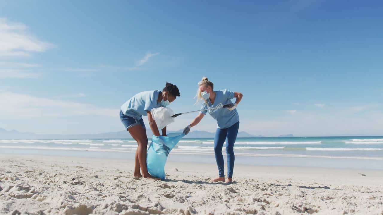 두 명의 다양한 여성이 자원봉사자 티셔츠와 얼굴 마스크를 입고 해변에서 쓰레기를 모으고 있습니다.