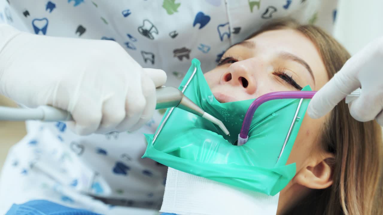 The dentist using the device rinses the tooth with water during the tooth treatment procedure. The girl in the dentist's office with open mouth