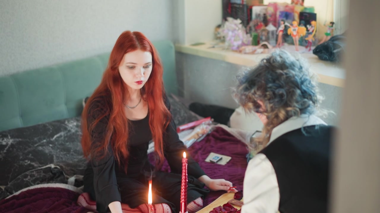 Lys, tarotlesing, kaukasisk spåkone, eldre krøllhåret leser lener seg over et stearinlysbelyst klede mens rødhåret klient sitter på sengen, kort spredt ut, røyk og stillhet, anspent forventning i bohemsk stil.