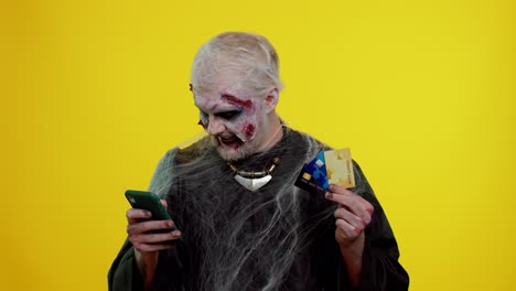 Sinister-man-Halloween-zombie-using-credit-bank-cards-and-smartphone-while-purchases-online-shopping