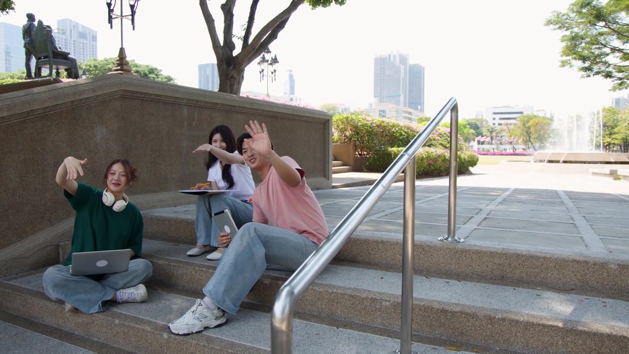 Group of college friends uses digital devices and waves while sitting on outdoor campus stairs.