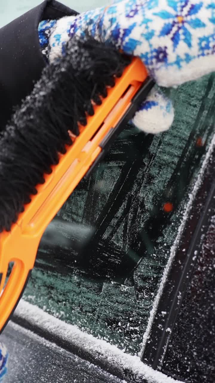 Uma mão com luva usa um raspador para remover gelo e neve da janela lateral de um carro, restaurando a visibilidade em uma manhã fria de inverno. Segurança no trajeto e material de apoio sobre o clima