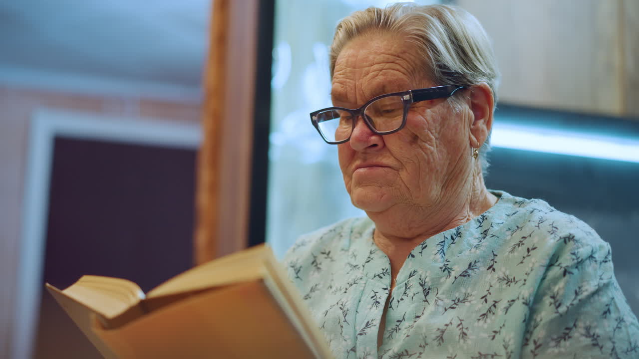 Donna anziana con occhiali assorta in un libro. Nonna anziana che si concentra serenamente sul suo libro aperto. Anziana nonna immersa nella lettura in un ambiente domestico tranquillo.