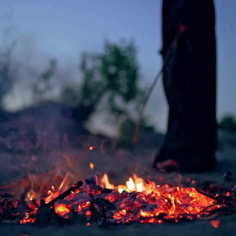 scary witch evil with a stick near the fire. dark death next to smoldered logs of fire in the evening. horror atmosphere outdoors.
