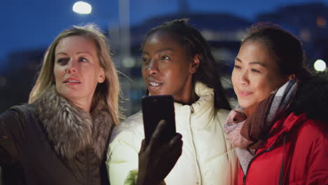 grupo de amigas en la calle de la ciudad por la noche pidiendo un taxi usando una aplicación de teléfono móvil