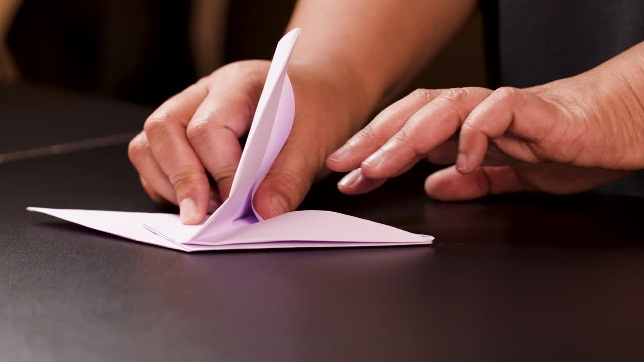 Adult hands carefully fold white paper into an origami boat, close-up, warm indoor lighting