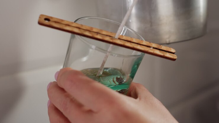Close-up of individual pouring greenish-blue liquid into glass cup with wick centered, wooden sticks balancing it, the liquid fills the cup for candle making process