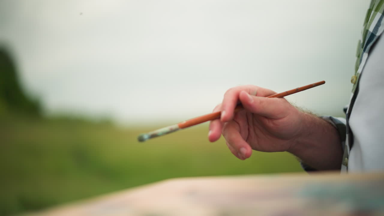 A close-up shot of an artist's hand delicately holding a paintbrush, with a blurred palette in the background. The serene, natural setting adds to the creative atmosphere
