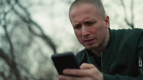 a close view of a man intently looking at his smartphone, displaying a serious expression, he is outdoors with a blurred background of trees