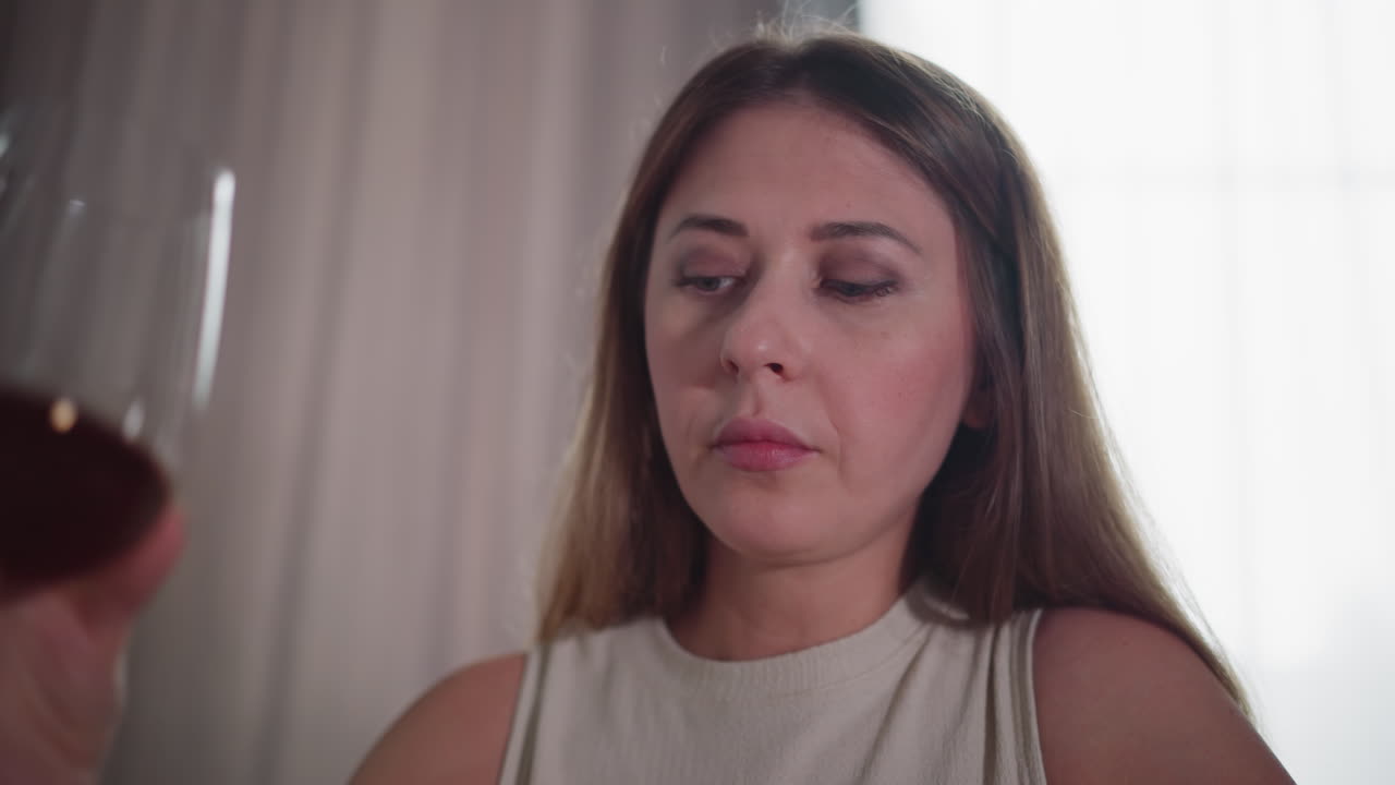 Close up of young woman in sleeveless outfit drinking red wine quickly from large glass, head tilted back, natural indoor lighting with soft curtain background