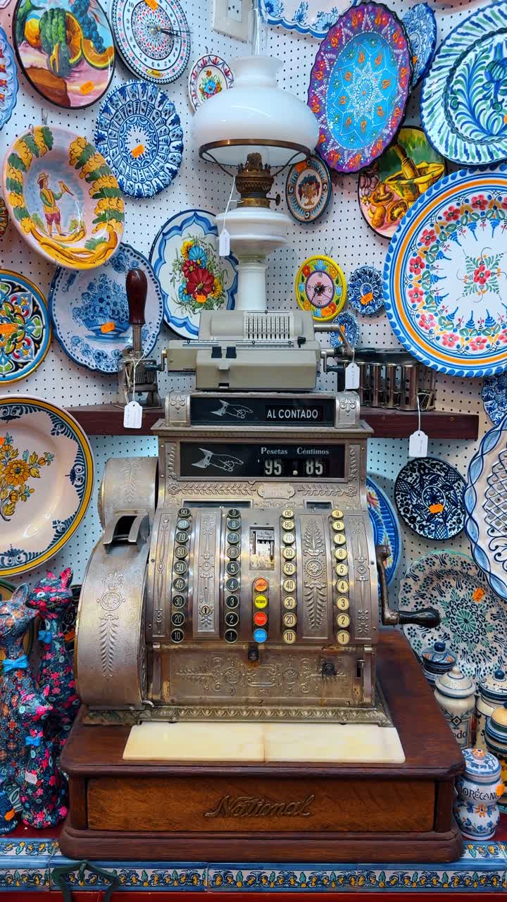 Alicante, Spagna, 6 agosto 2025: Registratore di cassa meccanico d'epoca ed esposizione di piatti di ceramica colorati