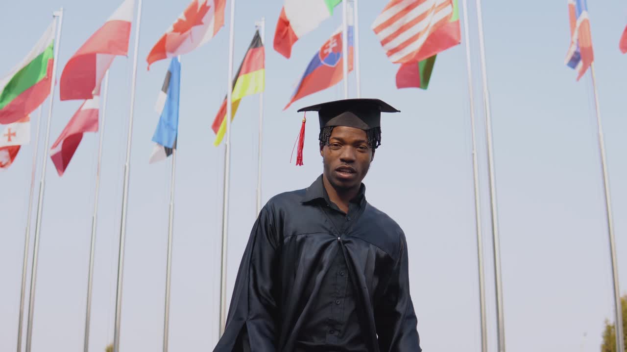 Very young african american male graduate standing in front of the camera with a diploma in his hands. The student hat and stands outside with the international flags on background.