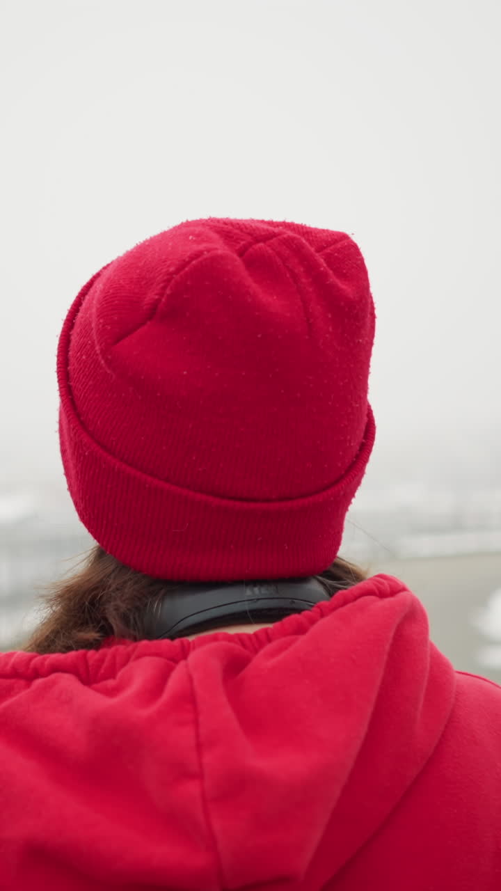 Close-up back view of woman in red beanie and jacket gazing upward with headphones around neck, surrounded by snow-covered environment with iron railing and blurred cityscape in foggy winter