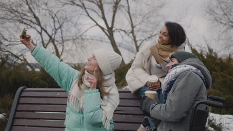 grupo de amigos, dos mujeres y un hombre discapacitado en silla de ruedas, tomando un video selfie en un parque urbano durante la temporada de invierno