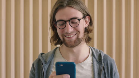 Primer-Plano-Retrato-De-Un-Lindo-Hombre-Geek-Con-Gafas-Sonriendo-Disfrutando-De-La-Navegación-De-Mensajes-De-Texto-Usando-Tecnología-Móvil-De-Teléfono-Inteligente-En-El-Interior