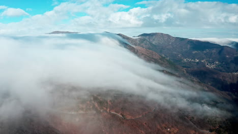 Clouds-rolling-over-mountain-peaks-with-clear-blue-sky,-aerial-view