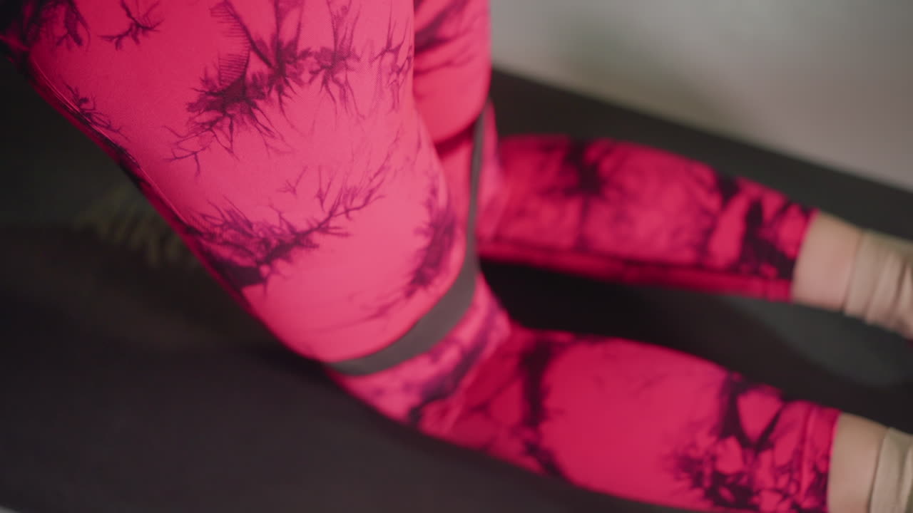 Atleta donna in leggings rosa inginocchiata su tappetino nero che esegue una lenta attivazione dei glutei, primo piano di leggings fantasia, luce soffusa dello studio, respirazione controllata, riscaldamento a basso impatto per la mobilità dell'anca