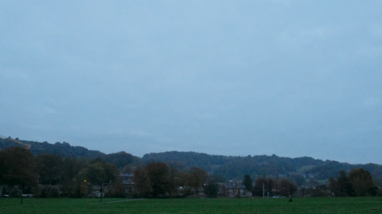 A man walks his black dog on the grass in a park, shot first thing in the morning before sunrise