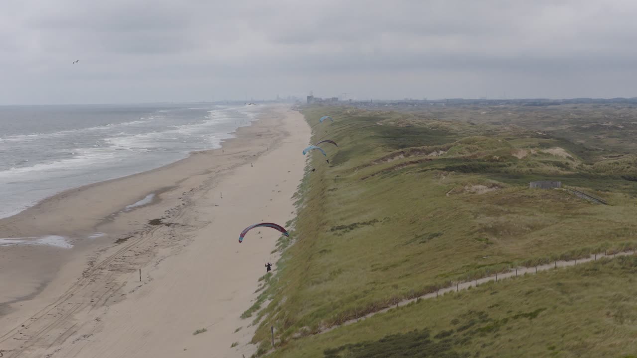 Una cámara profesional se desplaza de izquierda a derecha, creando un efecto de paralaje sobre las dunas de Langevelderslag. Una gaviota cruza el encuadre mientras parapentes se elevan sobre la playa del Mar del Norte en la distancia.