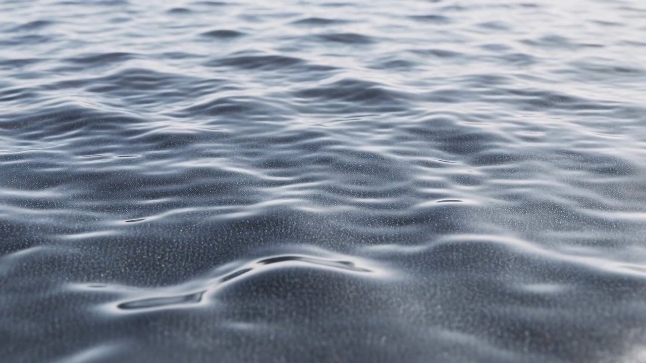 Low near-waterline view of calm open ocean surface showing small ripples, shallow undulations and soft reflections across wide water texture in cool blue gray tones, with water, sea, ocean, surface